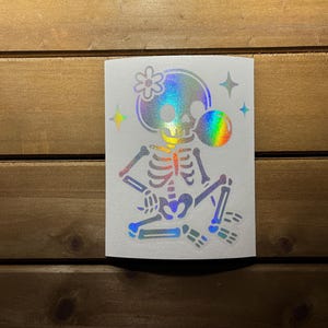 May include: A holographic sticker featuring a skeleton with a flower on its head and a rainbow bubble. The skeleton is in a relaxed pose, with stars on either side. The sticker has a white background.