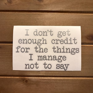 May include: A white rectangular sign with the text "I don't get enough credit for the things I manage not to say" in a typewriter font. The sign is set against a brown wooden background.