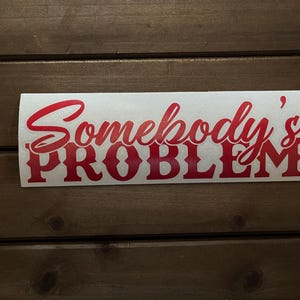 May include: A white sticker with the words "Somebody's PROBLEM" in red script and block letters. The sticker is rectangular and has rounded corners. The background is a dark brown wooden surface.