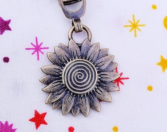 May include: Antique gold-toned zipper pull with a sunflower design. The sunflower has a spiral in the center and is surrounded by petals.
