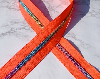 Puede incluir: Una cinta de cremallera de color naranja brillante con una cadena de cremallera iridiscente arcoíris. La cinta de cremallera está doblada por la mitad y la cadena de cremallera es visible en ambos lados.