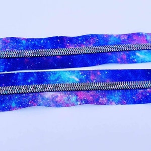 Puede incluir: Dos cintas de cremallera con estampado de galaxia. La cinta es azul con detalles en rosa y blanco. Los dientes de la cremallera son plateados.