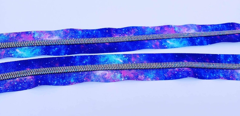 Puede incluir: Dos cintas de cremallera con estampado de galaxia. La cinta es azul con detalles en rosa y blanco. Los dientes de la cremallera son plateados.