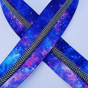 Puede incluir: Una cremallera con estampado de galaxia. La cremallera es azul, rosa y blanca con un tirador de cremallera plateado.