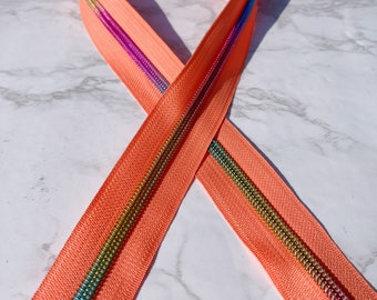 Puede incluir: Primer plano de una cremallera de color coral con una cinta de cremallera iridiscente arcoíris. La cremallera está dispuesta en forma de ocho.