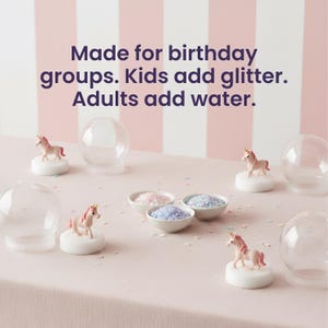 May include: A pink and white striped backdrop with a light pink tablecloth. The image shows clear glass globes, small unicorn figurines, and bowls of glitter. Text reads: Made for birthday groups. Kids add glitter. Adults add water.