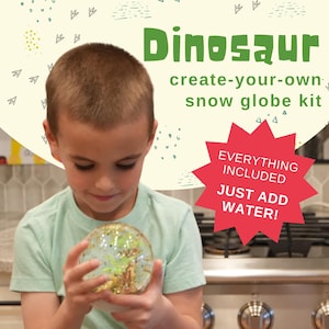 May include: A child holds a clear snow globe with green and gold glitter inside. The snow globe is part of a create-your-own dinosaur snow globe kit. The text on the background reads "Dinosaur create-your-own snow globe kit" and "Everything Included Just Add Water!"