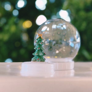 May include: A miniature Christmas tree figurine inside a clear glass snow globe with a white base. The snow globe is sitting on a white surface.