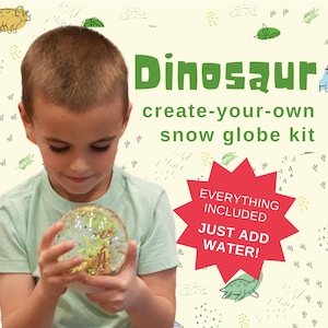 May include: A child holds a clear snow globe with a green and gold dinosaur inside. The snow globe is part of a create-your-own snow globe kit. The text on the image reads "Dinosaur create-your-own snow globe kit. Everything included. Just add water!"
