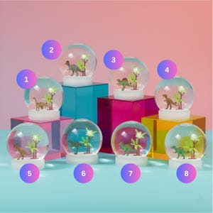 May include: A collection of eight dinosaur-themed snow globes. Each globe features a miniature dinosaur figure and artificial plants inside a clear, spherical glass enclosure. The globes sit on colorful, geometric display blocks.
