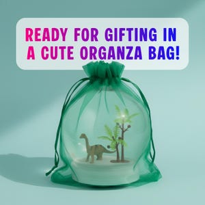 May include: A clear globe with a dinosaur and palm tree figurines inside, presented in a green organza bag. The text "READY FOR GIFTING IN A CUTE ORGANZA BAG!" is displayed above the bag against a teal background.