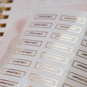 以下が含まれることがあります： 「APPOINTMENT」と金色の文字が書かれた白いステッカーのシートです。