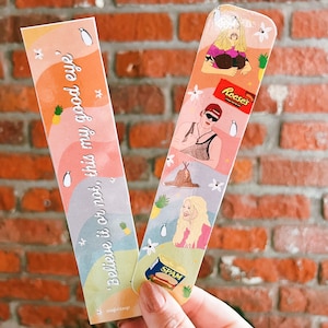May include: Two colorful bookmarks with illustrations and text. One bookmark has the text "Believe it or not, this my good eye" with a penguin and pineapple graphic. The other bookmark has illustrations of people, a Reese's candy bar, and a can of Spam.