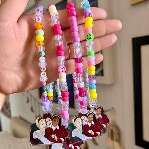 May include: Handmade beaded keychains in various colors, including pink, yellow, and clear beads. Each keychain features a charm with a group of three women. Some beads spell out names and feature smiley faces.