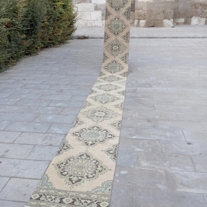 Pode incluir: Um longo tapete corredor retangular bege com um intrincado padrão floral em tons de castanho, verde e azul. O tapete está enrolado em uma extremidade e repousa sobre uma superfície de pedra, mostrando seu design detalhado.