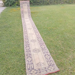 Op de afbeelding: Een lang, smal loperkleed met een beige middenveld met herhalende donkergrijze geometrische bloemmotieven. De rand toont een complex donkergrijs patroon op een beige achtergrond, afgewerkt met een dunne rode rand aan de uiteinden.
