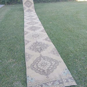 Può includere: Un tappeto passatoia lungo e stretto caratterizzato da una base color beige con motivi geometrici a medaglione marrone scuro ripetuti. Il tappeto presenta dettagli decorativi sui bordi con sottili accenti azzurri e grigi.