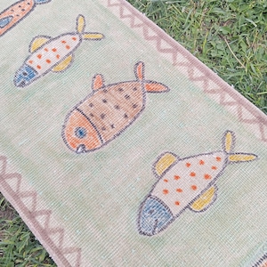May include: A light green runner rug featuring a repeating pattern of stylized fish in orange, yellow, and blue hues. The rug is bordered by a brown zigzag geometric design along the edges, creating a distinct frame for the central fish motifs.