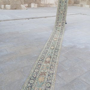 May include: A long, rectangular rug with an intricate pattern of beige, brown, and teal geometric and floral designs. The rug is displayed outdoors, leaning against a stone wall. The rug's design features a central medallion and border details.