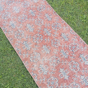 May include: A long, rectangular runner rug featuring a faded terracotta orange base color with a repeating pattern of stylized blue floral motifs and interconnected vine-like lines woven throughout the entire surface of the textile piece.
