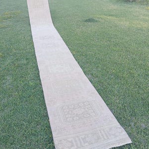 May include: A long, narrow runner rug in a light beige or cream color. The rug features a repeating pattern of geometric and floral motifs arranged in square sections along its length, with a decorative border at the end of the runner.