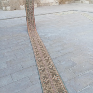 Peut inclure: Un long tapis étroit avec un motif géométrique détaillé dans des tons de marron, de rose et de turquoise. Le tapis est déroulé sur une surface en pierre, mettant en valeur son design et sa texture complexes. Les bords du tapis sont finis avec une frange.