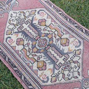 May include: A rectangular woven rug featuring a dusty pink background with a central geometric medallion pattern in shades of cream, slate blue, and mustard yellow. The border displays a dark grey intricate design with small floral motifs.