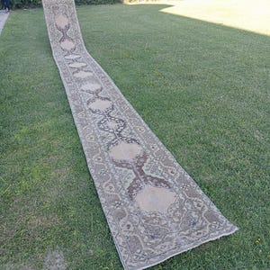 May include: An exceptionally long, narrow runner rug featuring a repeating geometric pattern in muted shades of beige, brown, and charcoal grey. The rug displays a series of interconnected medallions and intricate border details throughout its length.