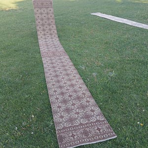 May include: A long, rectangular rug with a detailed floral pattern in shades of brown and beige. The rug is displayed outdoors on green grass, showcasing its length and intricate design. The rug's edges have a decorative border.