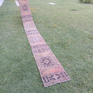 Puede incluir: Una alfombra de pasillo larga y estrecha con un patrón repetitivo de motivos geométricos y florales de color marrón oscuro sobre un fondo de color tostado claro descolorido. La alfombra tiene forma rectangular y textura plana.