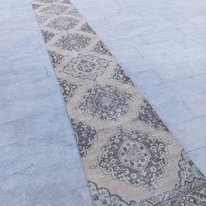 Pode incluir: Um tapete longo e retangular com um padrão de medalhão repetido em tons de bege, castanho e azul suave. O tapete tem uma aparência desgastada e vintage, adequado para um corredor ou entrada. O design do tapete apresenta elementos florais e geométricos.
