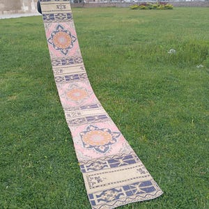 Pode incluir: Um tapete longo e retangular com um padrão floral em rosa, bege e azul marinho. O tapete é exibido ao ar livre sobre grama verde. O design floral é centrado com uma flor cor de pêssego.