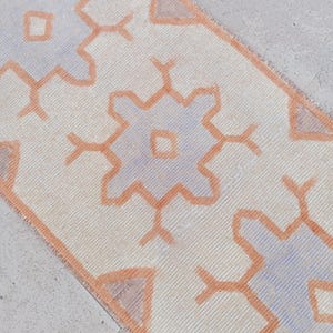 Puede incluir: Una alfombra rectangular de color crema con un patrón geométrico repetitivo en naranja claro y azul pálido. El diseño de la alfombra presenta formas estilizadas de flores o estrellas, creando una apariencia simétrica y decorativa. La alfombra es probablemente de material tejido.