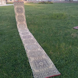 Puede incluir: Una alfombra rectangular y larga con un patrón geométrico repetitivo en tonos beige, gris y rojo. La alfombra está desenrollada sobre una superficie cubierta de hierba, mostrando su intrincado diseño y textura. Los bordes de la alfombra están acabados con un borde rojo.