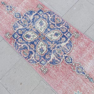 May include: A rectangular, distressed-look rug with a pink background and a large, ornate blue and white central design. The rug features a detailed, symmetrical pattern with floral and geometric elements, suitable for a hallway or entryway.
