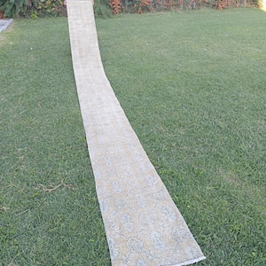 May include: A long, narrow runner rug featuring a muted beige and light blue floral pattern. The rug has a rectangular shape with short white fringe detailing at the bottom edge, displaying a repeating botanical design across its entire surface.