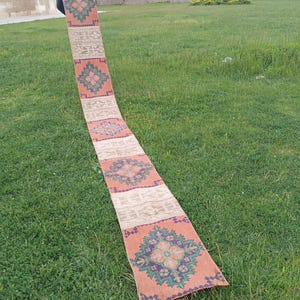 May include: A long, narrow rug with a repeating geometric pattern in orange, green, and beige. The rug is displayed on grass. The design features floral and geometric motifs. The rug's length is suitable for a hallway or runner.