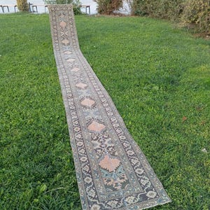 May include: A long, narrow runner rug featuring a repeating geometric pattern in muted shades of peach, sage green, and charcoal grey. The textile displays a central row of diamond-shaped medallions framed by intricate floral and vine-like borders.