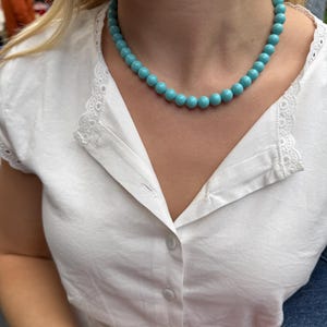 Puede incluir: Un collar de cuentas turquesas. El collar est&aacute; compuesto por cuentas redondas de color azul claro. El collar se lleva con una blusa blanca con detalles de encaje. Se ve una pulsera dorada en la mu&ntilde;eca.