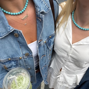 Puede incluir: Dos personas con collares de cuentas turquesas y chaquetas vaqueras. Una persona lleva una camiseta blanca, y la otra lleva una camiseta blanca con un collar de oro que dice "TOWN". Una persona sostiene un vaso de pl&aacute;stico transparente con una bebida verde.