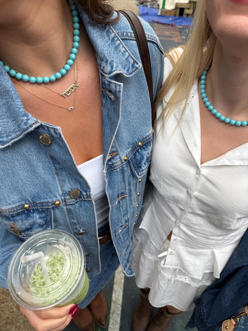 Puede incluir: Dos personas con collares de cuentas turquesas y chaquetas vaqueras. Una persona lleva una camiseta blanca, y la otra lleva una camiseta blanca con un collar de oro que dice "TOWN". Una persona sostiene un vaso de pl&aacute;stico transparente con una bebida verde.