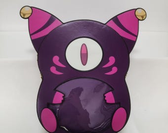 May include: A purple and pink cat-shaped bag with a clear front pocket. The bag features a large oval eye and decorative swirls. The ears are pink with gold accents. The bag is designed with a cartoonish aesthetic.