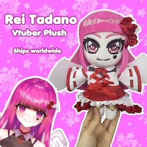 Puede incluir: Un juguete de peluche rosa y blanco de un personaje con cabello rosa, ojos rojos y un vestido rojo y blanco. El juguete tiene alas y un lazo rojo. El texto "Rei Tadano Vtuber Plush Ships worldwide" es visible en la imagen.