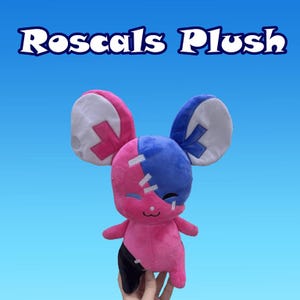 Peut inclure: Un jouet en peluche en forme de souris rose et bleu avec un visage cousu et des accents noirs. Le jouet a de grandes oreilles avec des patchs blancs et une croix cousue sur chaque oreille.