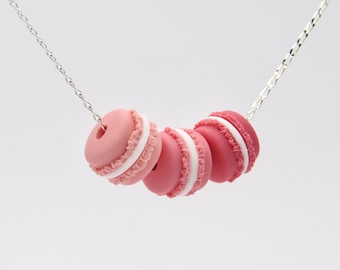 May include: A silver chain necklace with four pink and white macarons attached. The macarons are arranged in a gradient from light pink to dark pink.