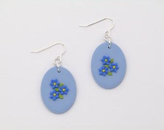 May include: A pair of oval-shaped earrings with a light blue background. Each earring features a cluster of blue flowers with yellow centers and green leaves. The earrings have silver-colored ear wires.