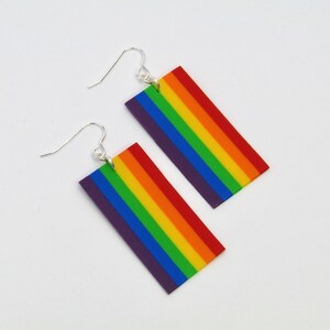 Peut inclure: Une paire de boucles d'oreilles pendantes arc-en-ciel. Les boucles d'oreilles sont en pâte polymère et présentent une forme rectangulaire avec six bandes de couleur : violet, bleu, vert, jaune, orange et rouge. Les boucles d'oreilles sont attachées à des crochets argentés.