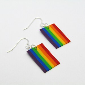 May include: A pair of rainbow flag earrings with silver hooks. The earrings are made of polymer clay and feature the colors of the rainbow flag: red, orange, yellow, green, blue, indigo, and violet.