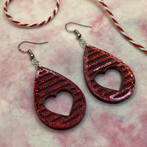 May include: A pair of red glitter teardrop earrings with a heart cut-out. The earrings have silver hooks.