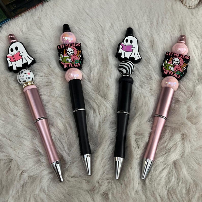 May include: Three pens with ghost and skeleton charms. The pens are black, pink, and rose gold. The charms say "Let me read in peace".